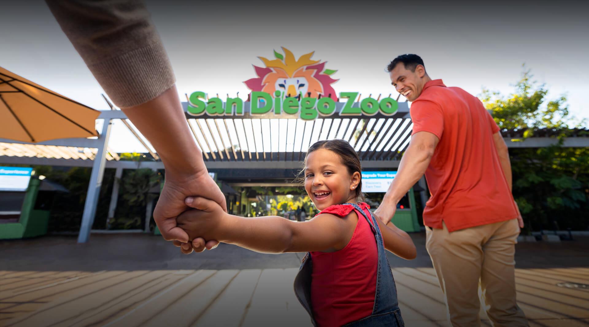 Girl holding parent's hands walking into the zoo